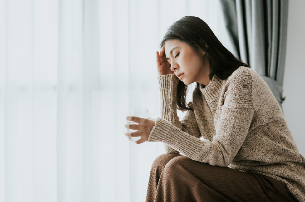 A side view of a young woman sitting down with her eyes closed, holding one hand to her forehead in pain or dizziness, indicating a migraine or stress. In her other hand, she holds a clear glass of water. She is wearing a cozy, beige textured knit sweater and is positioned next to a bright window with sheer white curtains.